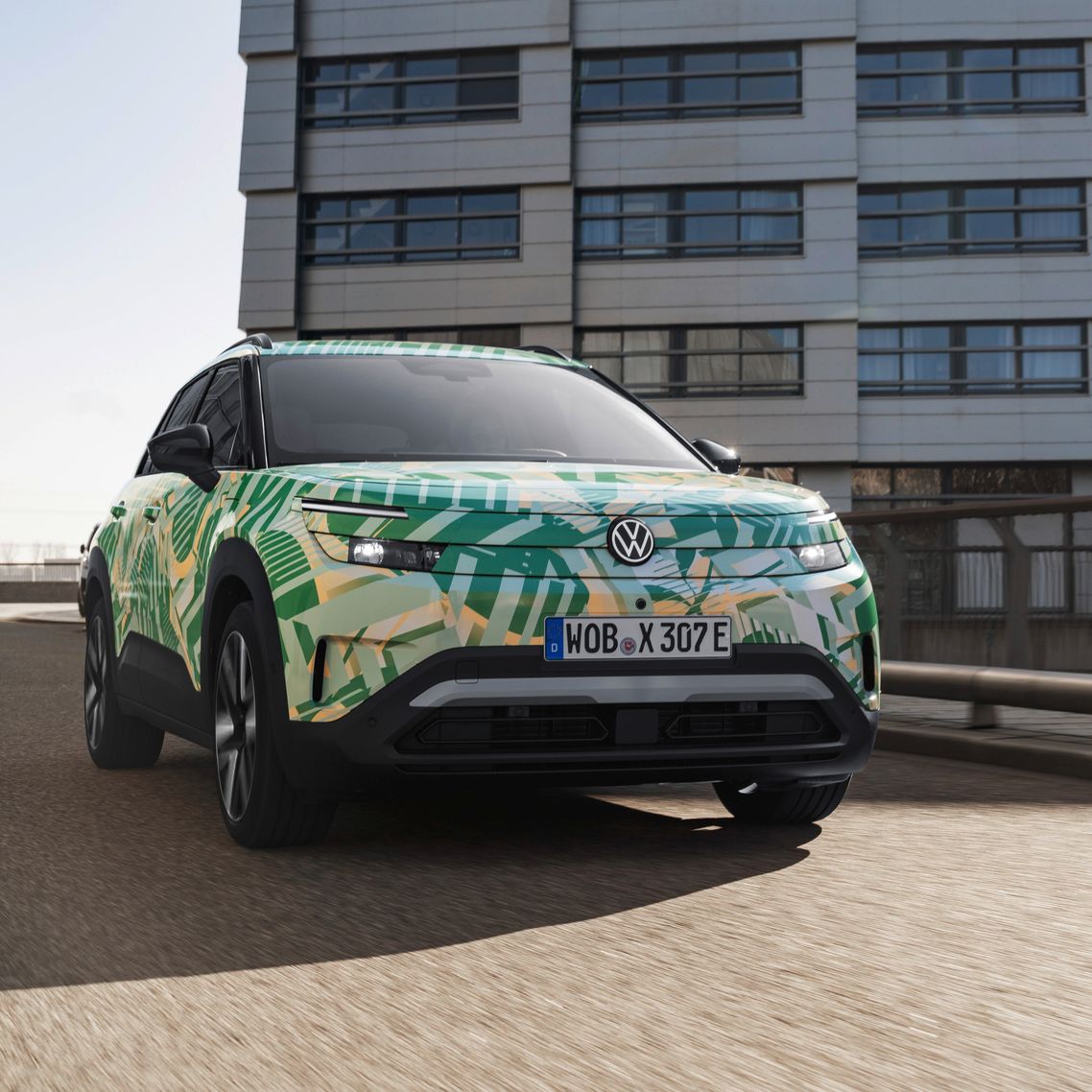 Ein ID. Cross von Volkswagen mit einem auffälligen grünen und gelben Camouflage-Design fährt auf einer städtischen Straße. Im Hintergrund sind moderne Gebäude zu sehen. Das Nummernschild des Autos beginnt mit "WOB".