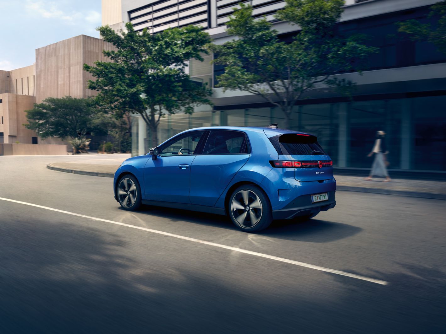Ein blauer VW ID. Polo Style fährt durch eine Stadt mit grünen Bäumen am Straßenrand.