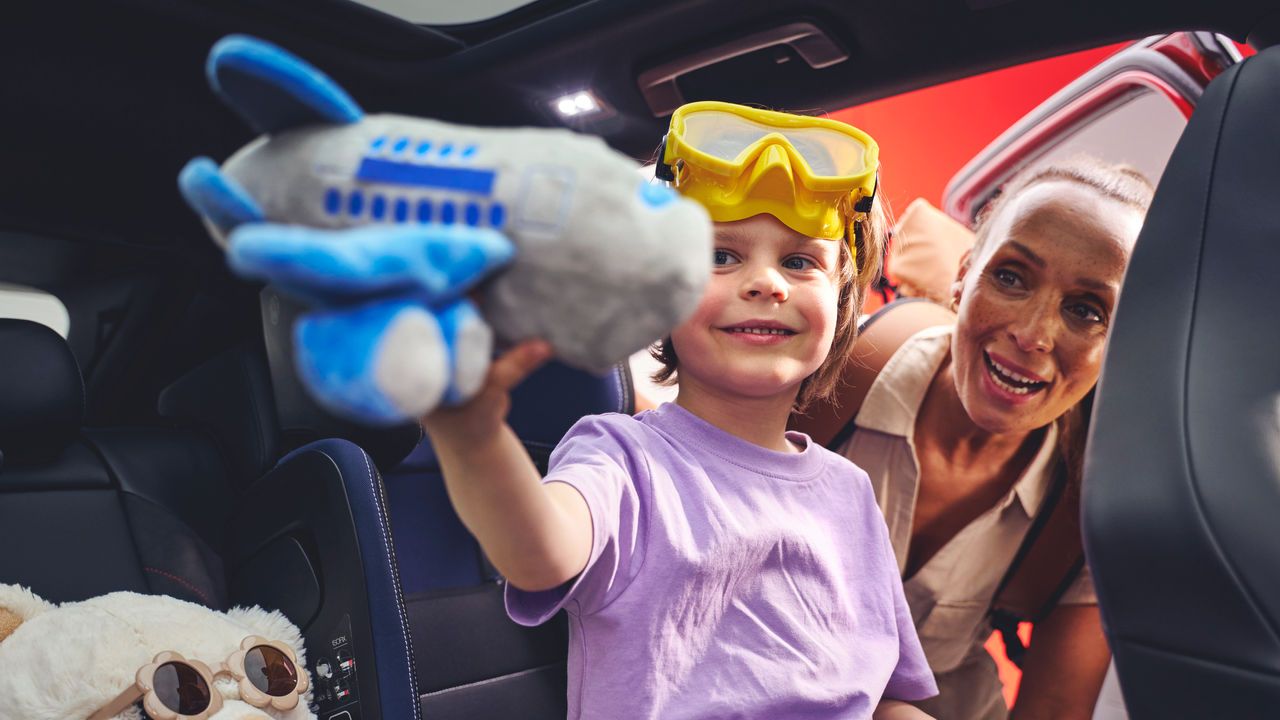Ein Kind in einem Volkswagen ID.4 hält ein Spielzeugflugzeug hoch. Das Kind trägt eine gelbe Schutzbrille. Neben dem Kind sitzt ein weißer Plüschbär mit Sonnenbrille, während eine andere Person im Hintergrund durch die Autotür zu sehen ist.