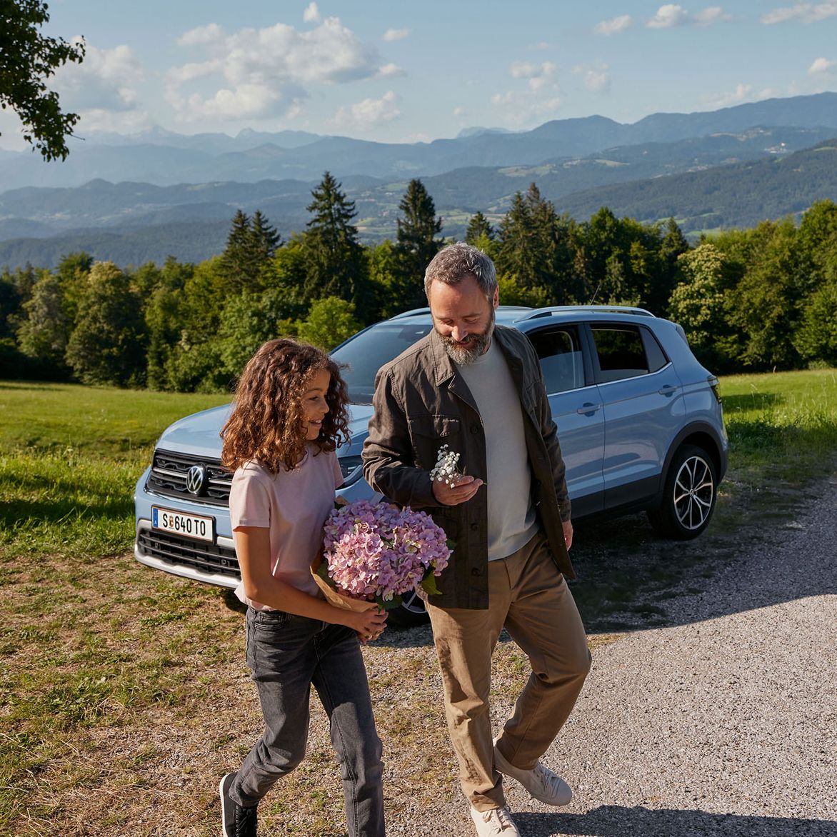 Ein blauer VW T-Cross steht geparkt auf einer Wiese in den Bergen. Davor laufen ein Mädchen und ein Mann mit Blumen in der Hand.