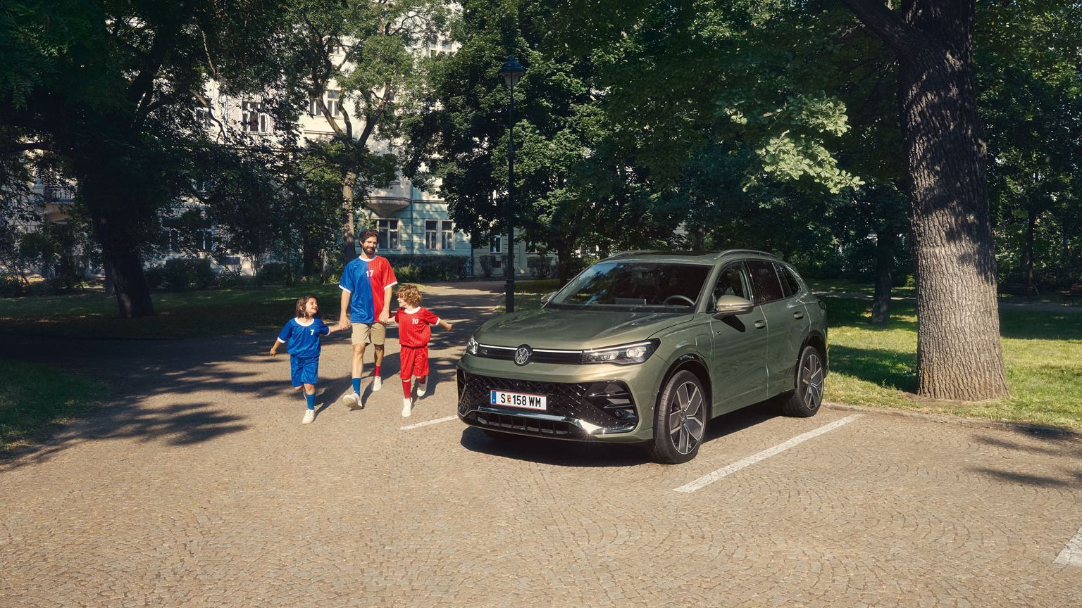 Ein Mann geht mit zwei Kindern von einem VW Tiguan R-Line Plug-In- Hybrid weg, der im Park steht.