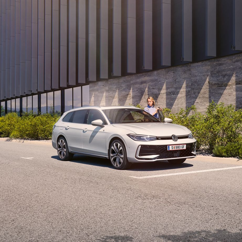  Eine Frau öffnet die Fahrertür eines weißen VW Passat R-Line und steigt ein. Im Hintergrund sieht man ein Gebäude.