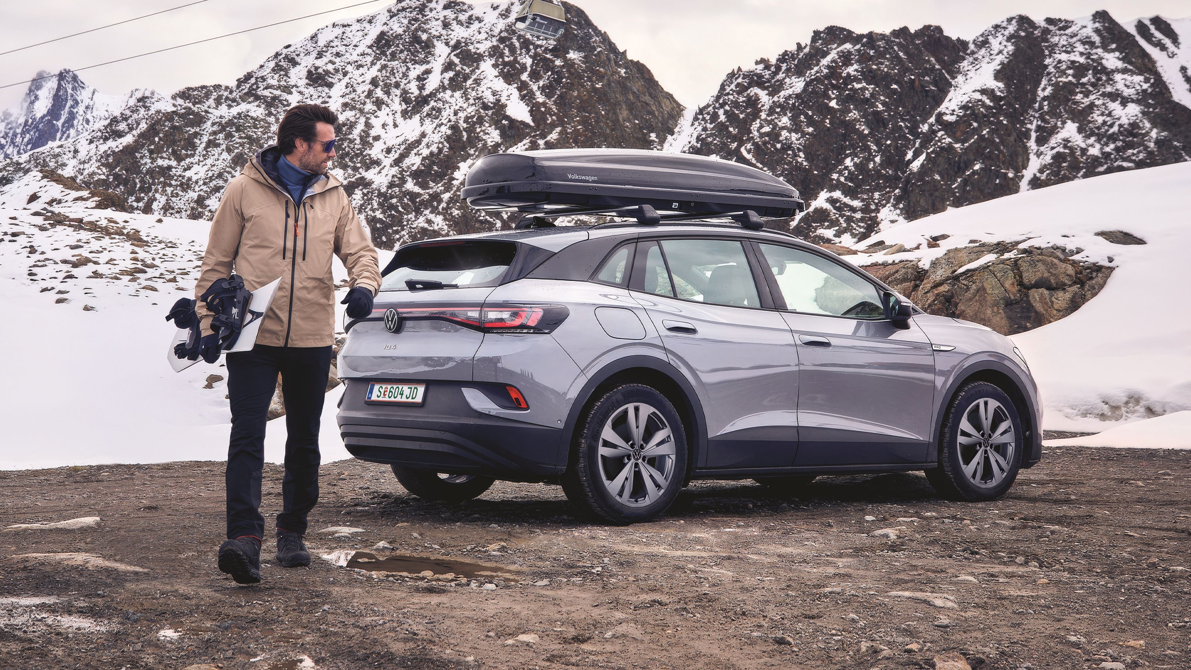 Ein Mann steht neben einem grauen Volkswagen SUV mit Dachbox. Er hält ein Snowboard in der Hand und trägt Winterkleidung, während der Boden matschig und teils von Pfützen bedeckt ist.