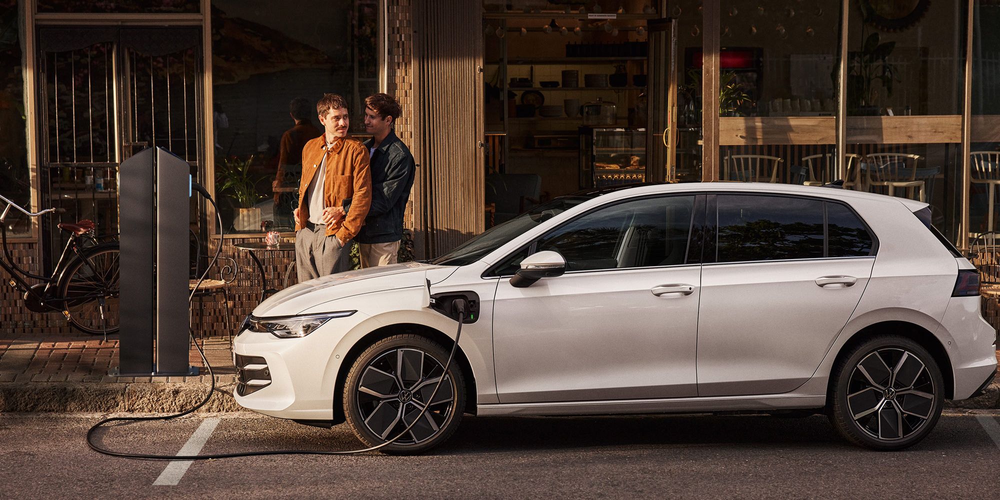 Ein weißer Golf eHybrid steht am Straßenrand und wird aufgeladen. Zwei Männer stehen neben dem Auto. 