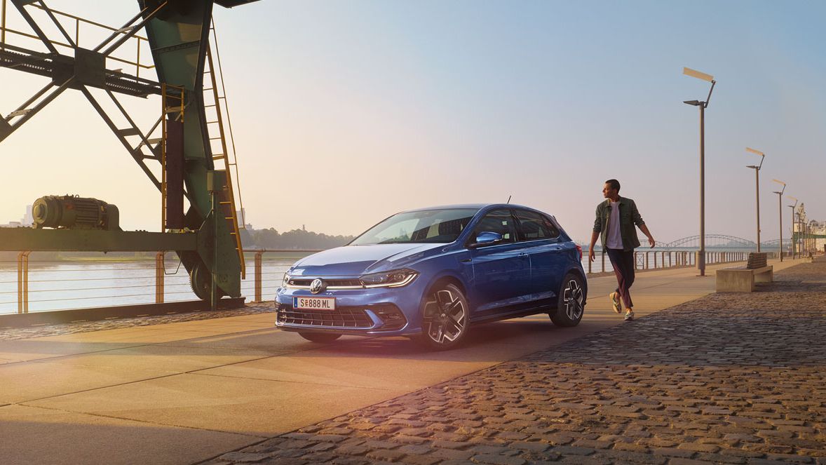  Ein blauer VW Polo R-Line steht an der Promenade neben einem Fluss, ein Mann läuft um das Auto herum.