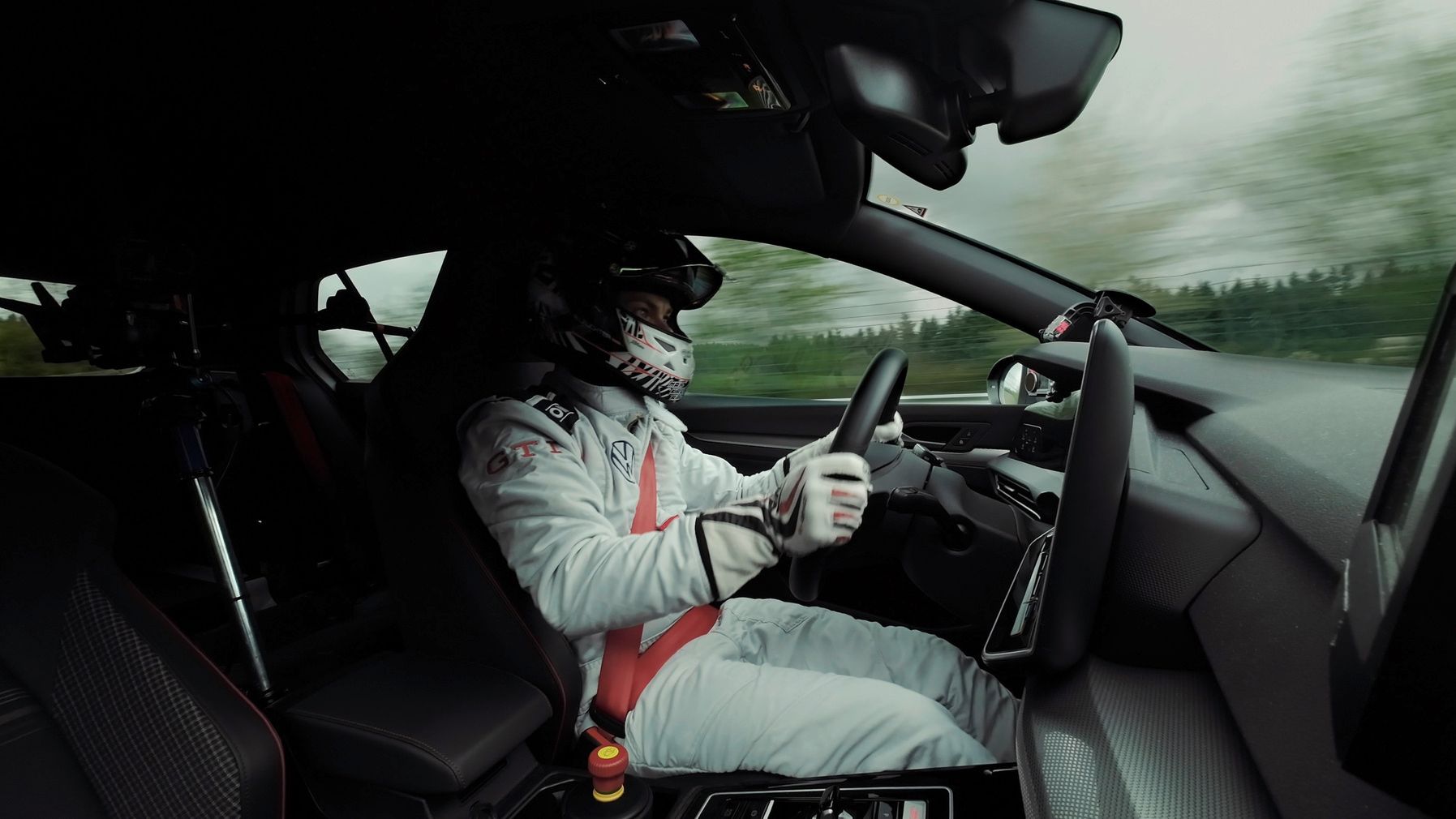 Ein Rennfahrer in voller Schutzkleidung sitzt im Cockpit eines Autos und blickt konzentriert auf die Rennstrecke. Der Innenraum des Autos ist minimalistisch und auf Funktionalität ausgelegt. Im Hintergrund sind schnell vorbeiziehende Bäume und Sträucher sichtbar.