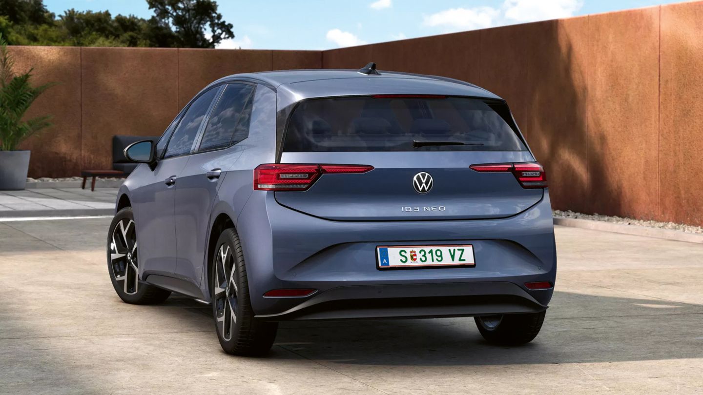 Ein grauer VW ID.3 Neo steht auf einem betonierten Platz, von hinten leicht schräg gesehen. Im Hintergrund sind eine niedrige, rostfarbene Mauer und einige Bäume unter blauem Himmel zu sehen. Das Auto wirkt modern und kompakt, mit schmalen Rückleuchten und Elektrofahrzeug-Design.