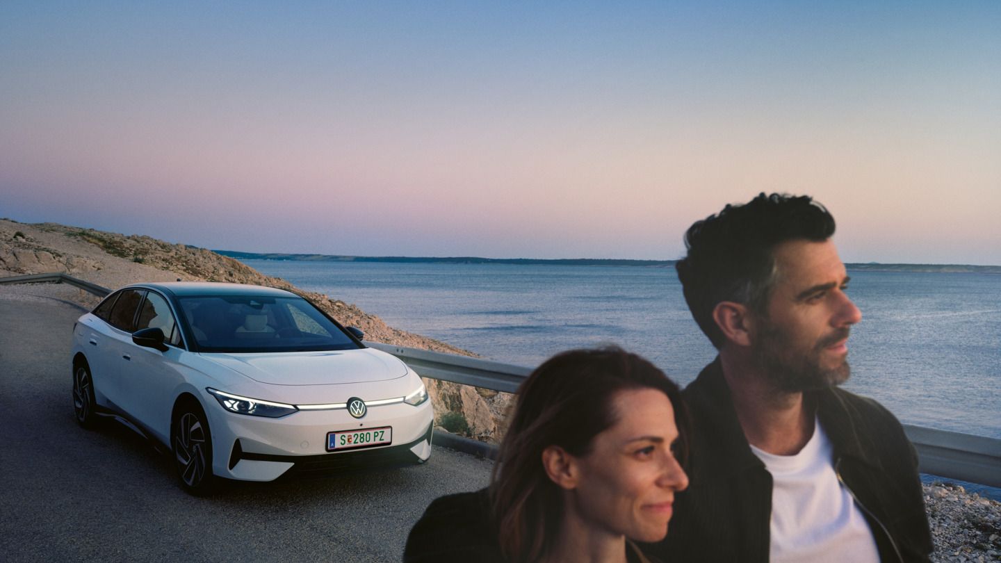 Ein Mann und eine Frau gehen Arm in Arm eine Straße am Meer entlang, im Hintergrund steht ein weißer VW ID.7.