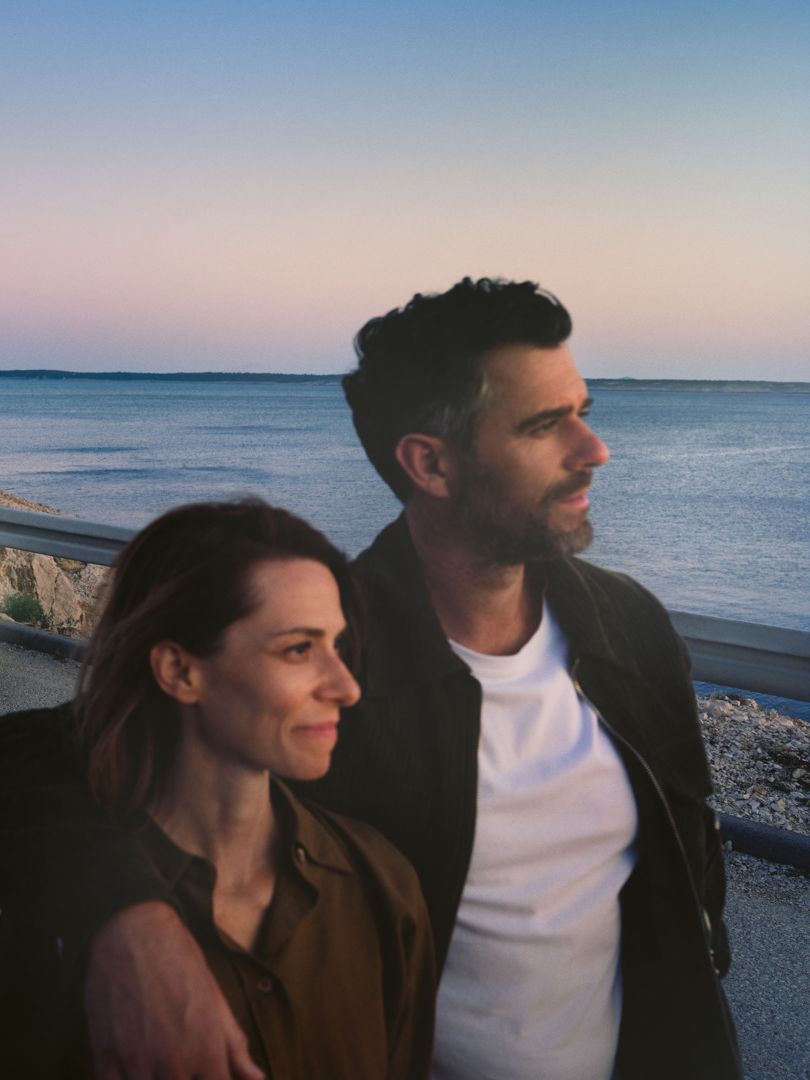 Ein Mann und eine Frau gehen Arm in Arm eine Straße am Meer entlang, im Hintergrund steht ein weißer VW ID.7.