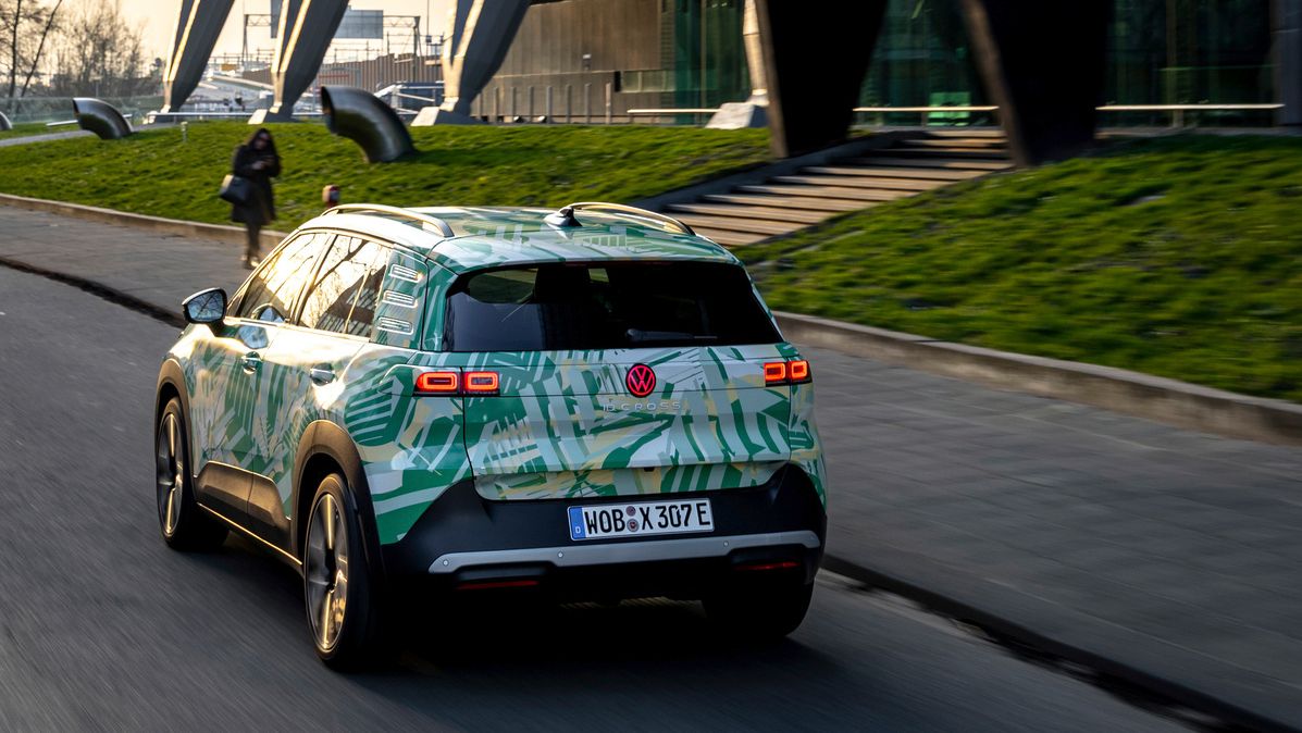 Ein Auto fährt auf einer Straße an einem modernen Gebäude mit Glasfassade und Stelzen vorbei. Das Auto hat eine auffällige, hellgrüne und gemusterte Lackierung. Im Hintergrund sind einige Bäume und eine Fußgängerin zu sehen.
