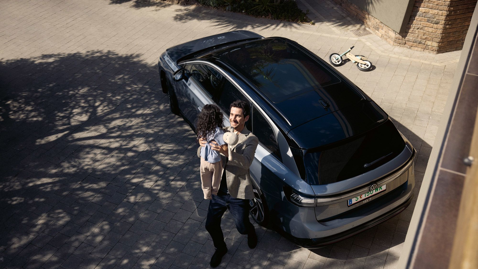 Mann hebt sein Kind hoch, was eine Puppe in der Hand halt und dahinter steht geparkt in der Einfahrt ein ID.7 Tourer und ein umgekipptes Kinderfahrrad.