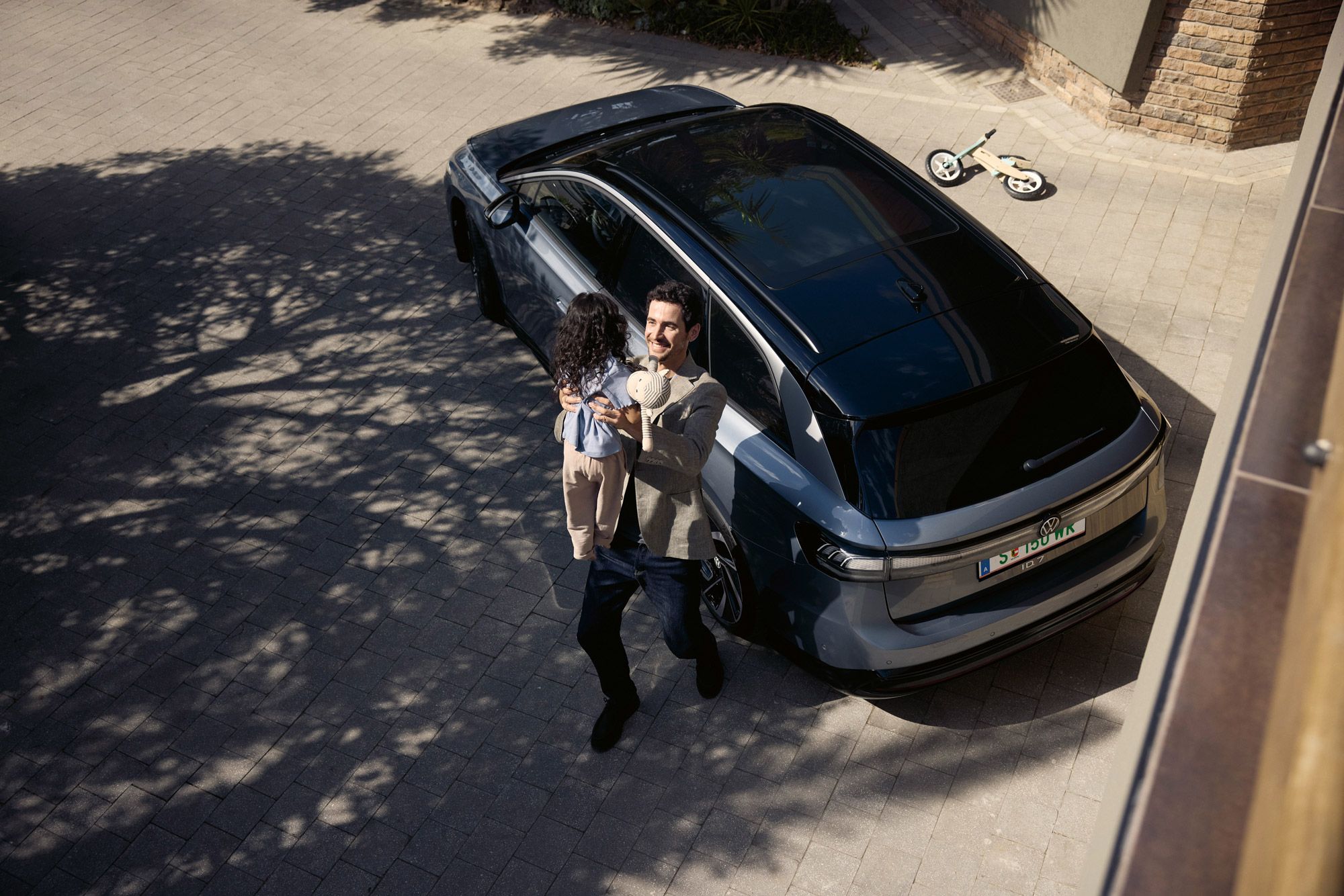 Mann hebt sein Kind hoch, was eine Puppe in der Hand halt und dahinter steht geparkt in der Einfahrt ein ID.7 Tourer und ein umgekipptes Kinderfahrrad.