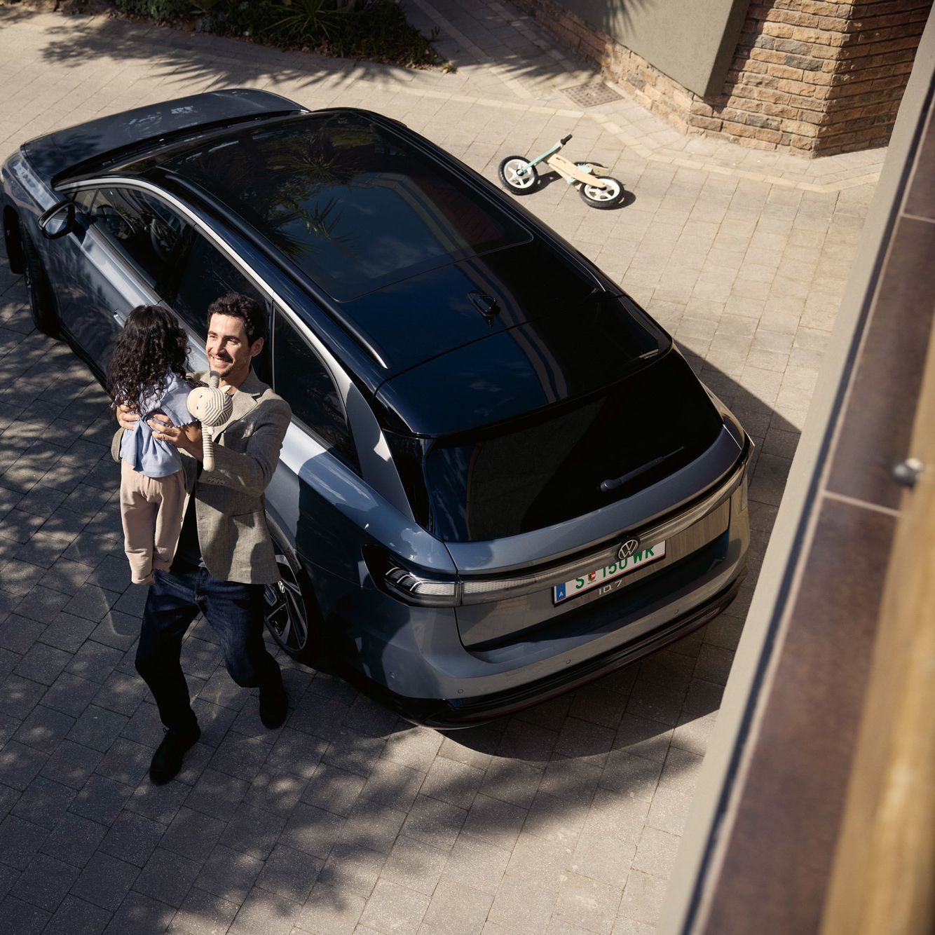 Mann hebt sein Kind hoch, was eine Puppe in der Hand halt und dahinter steht geparkt in der Einfahrt ein ID.7 Tourer und ein umgekipptes Kinderfahrrad.