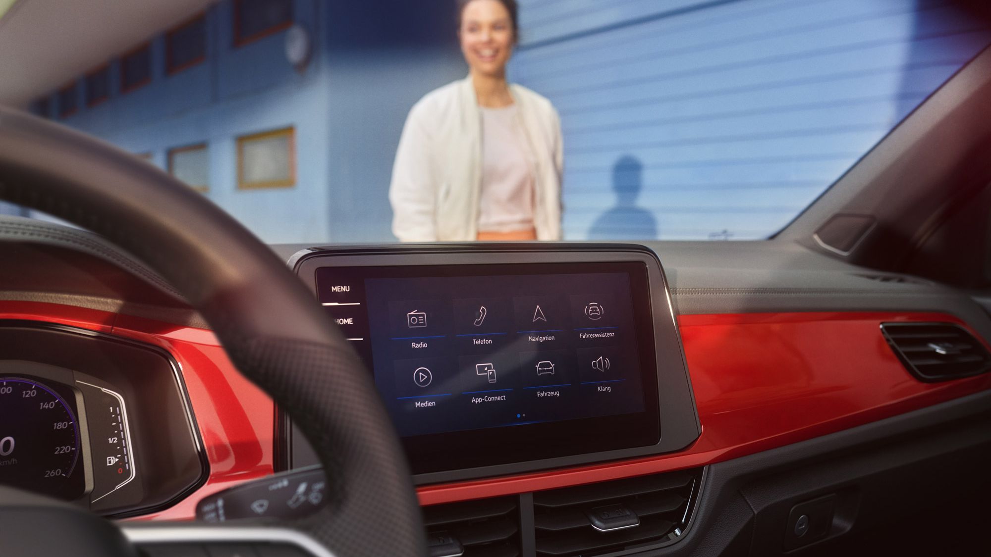 Das Bild zeigt das Innenleben eines VW T-Roc Cabrios mit einem modernen Infotainment-System. Im Hintergrund steht eine Person vor einer blauen Wand. Die Armaturenbrettdetails, wie rote Akzente, sind sichtbar.