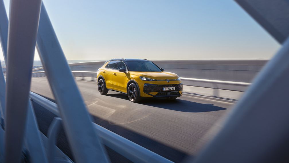 Ein gelber T-Roc fährt über eine moderne Brücke bei klarem Himmel. Die Aufnahme zeigt das Fahrzeug aus der Ferne, teilweise durch die Brückenstrukturen hindurch. Es ist ein sonniger Tag.