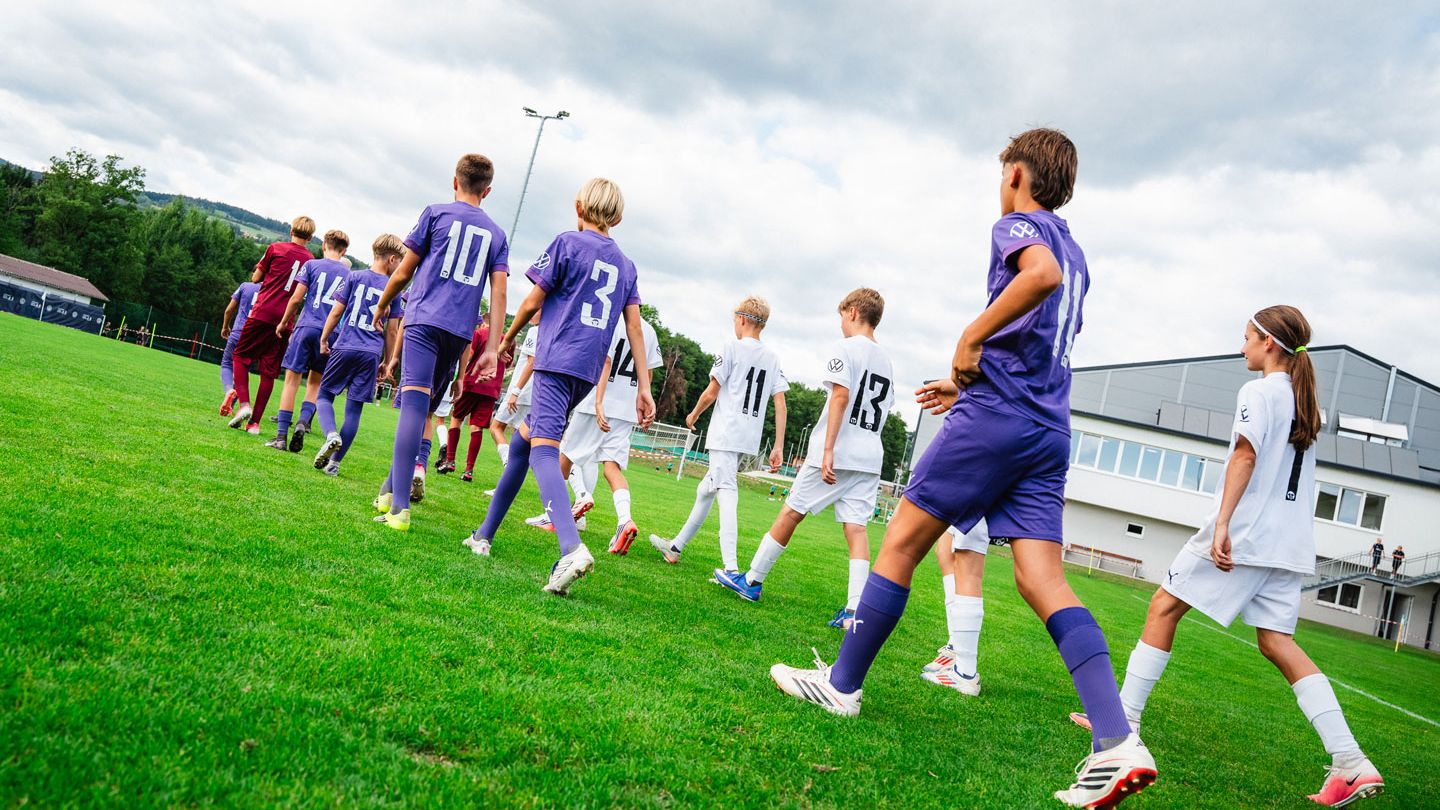 Mehrere Kinder-Fußballmannschaften laufen in Trikots auf ein grünes Spielfeld, die einen in Lila, die anderen in Weiß. Die Kinder sind von hinten zu sehen und bewegen sich in einer Reihe Richtung Spielfeldmitte. Im Hintergrund erkennt man ein Vereinsgebäude und Bäume unter einem bewölkten Himmel.