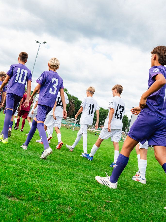 Mehrere Kinder-Fußballmannschaften laufen in Trikots auf ein grünes Spielfeld, die einen in Lila, die anderen in Weiß. Die Kinder sind von hinten zu sehen und bewegen sich in einer Reihe Richtung Spielfeldmitte. Im Hintergrund erkennt man ein Vereinsgebäude und Bäume unter einem bewölkten Himmel.