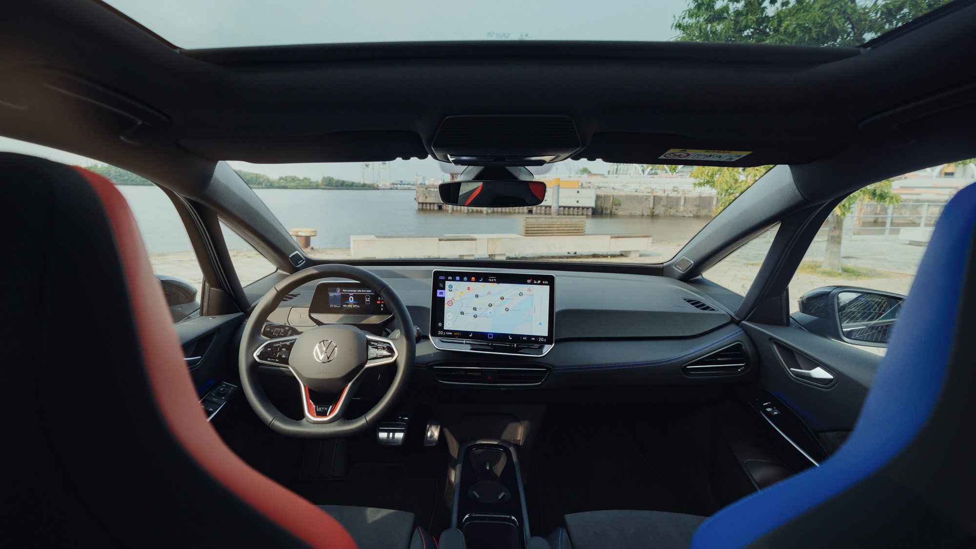 Innenraum des ID.3 GTX FIRE+ICE, das mit einem großen Touchscreen-Navigationsgerät ausgestattet ist. Zwei vordere Sitze, einer in Rot und einer in Blau, sind sichtbar. Die Aussicht aus dem Fenster zeigt eine Uferlandschaft.