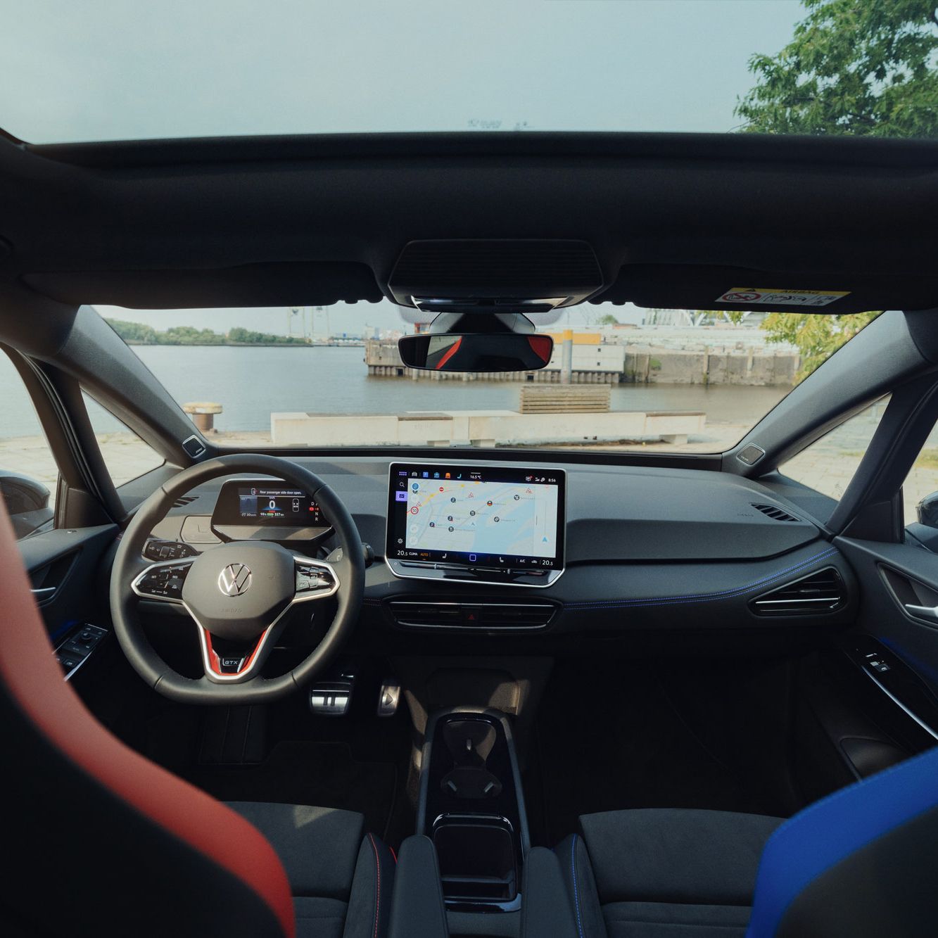 Innenraum des ID.3 GTX FIRE+ICE, das mit einem großen Touchscreen-Navigationsgerät ausgestattet ist. Zwei vordere Sitze, einer in Rot und einer in Blau, sind sichtbar. Die Aussicht aus dem Fenster zeigt eine Uferlandschaft.