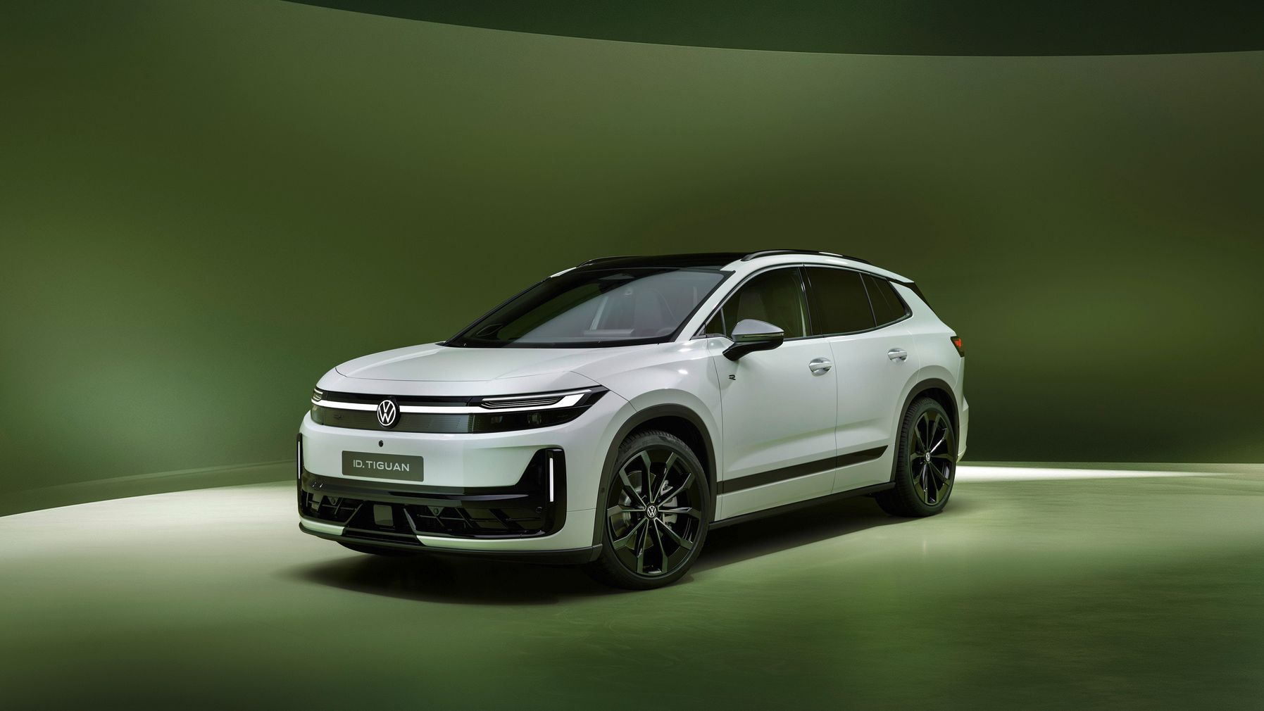 Ein weißer, moderner SUV steht leicht schräg von vorn auf einer glänzenden, grünen Studiobühne. Das Fahrzeug trägt ein Volkswagen-Logo und hat schlanke LED-Scheinwerfer sowie schwarze Designelemente, unter anderem am Dach und an den Felgen. Der Hintergrund ist abstrakt und weich ausgeleuchtet, wodurch der Wagen im Mittelpunkt steht.