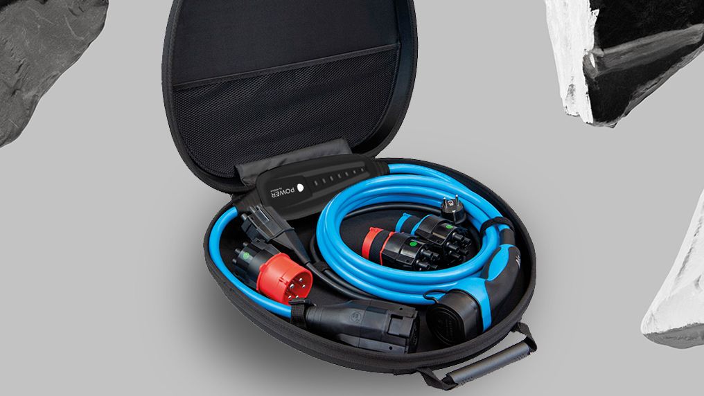 Ein offener, ovaler Koffer enthält blaue und schwarze Kabel sowie Anschlüsse. Der Hintergrund ist grau und mit schwarz-weißen Gesteinsformen verziert. Das Bild zeigt Ausrüstung für den Anschluß von Elektrofahrzeugen.