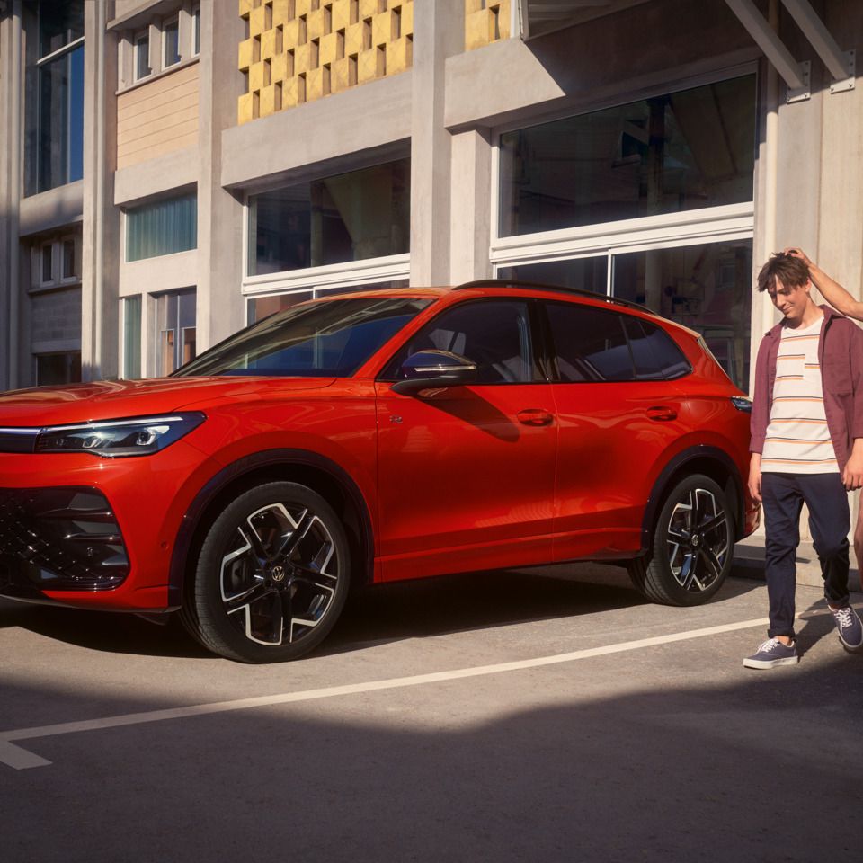 Eine Frau und ein Mann verlassen ein Gebäude mit großen Glasfenstern und gehen an einem roten VW Tiguan vorbei.