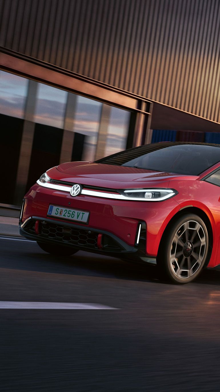 [AI-modified] – Ein roter VW ID.3 GTI fährt mit eingeschalteten Scheinwerfern und beleuchtetem Logo an einem Hafengebäude vorbei.