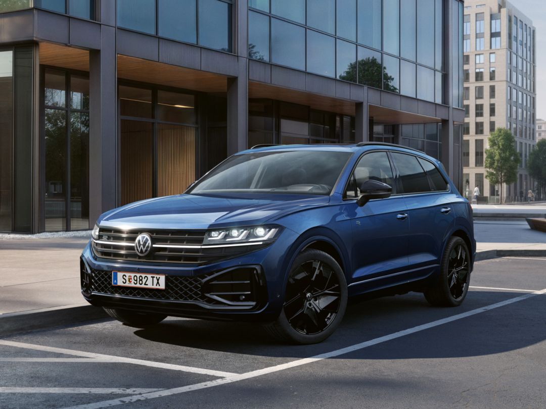 Ein blauer VW Touareg Final Edition steht vor einem Bürogebäude mit Glasfassade - Ansicht von schräg vorne.