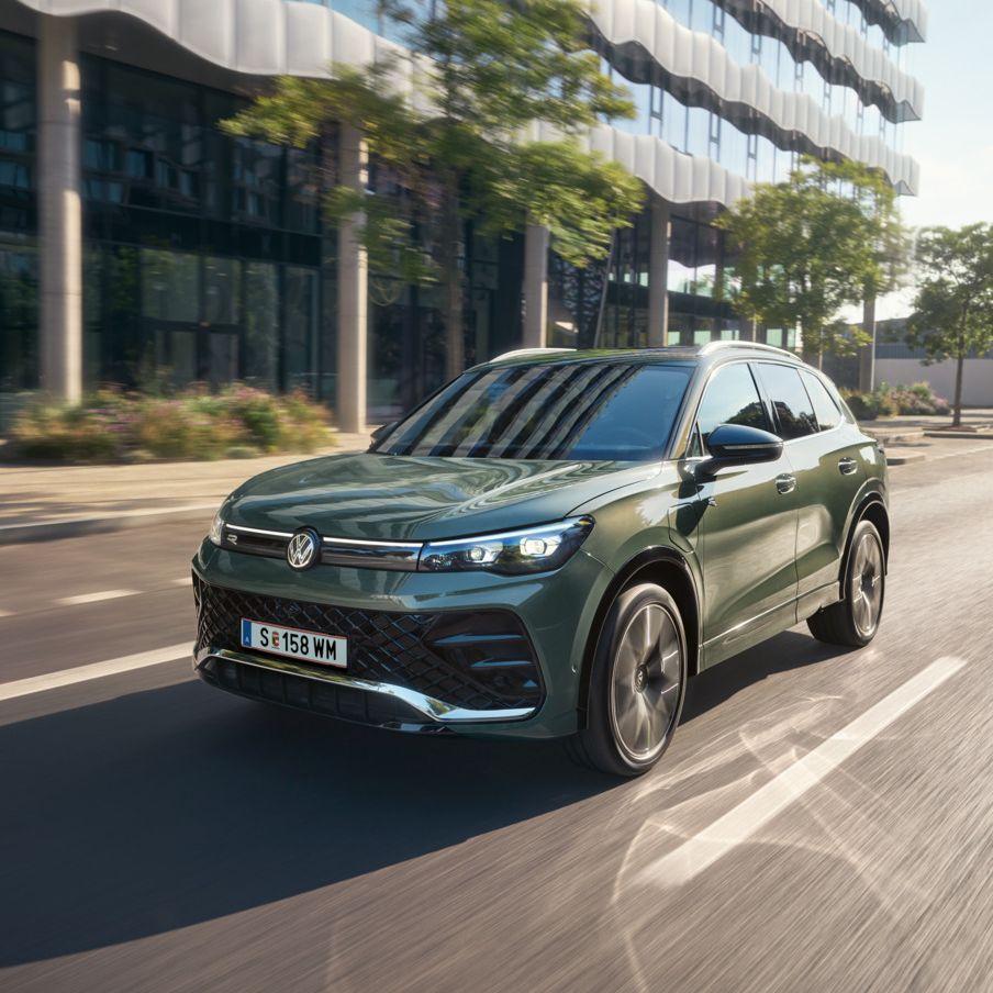 Ein Tiguan eHybrid in Cipresso-Grün fährt auf der Straße. Im Hintergrund sind ein modernes Gebäude und Bäume zu sehen. 