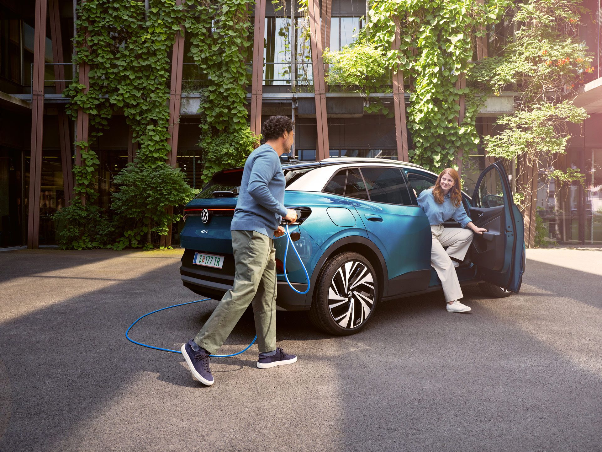 Ein Mann mit Ladekabel in der Hand geht zu einem blauen VW ID.4, der in einem begrünten Hof parkt.