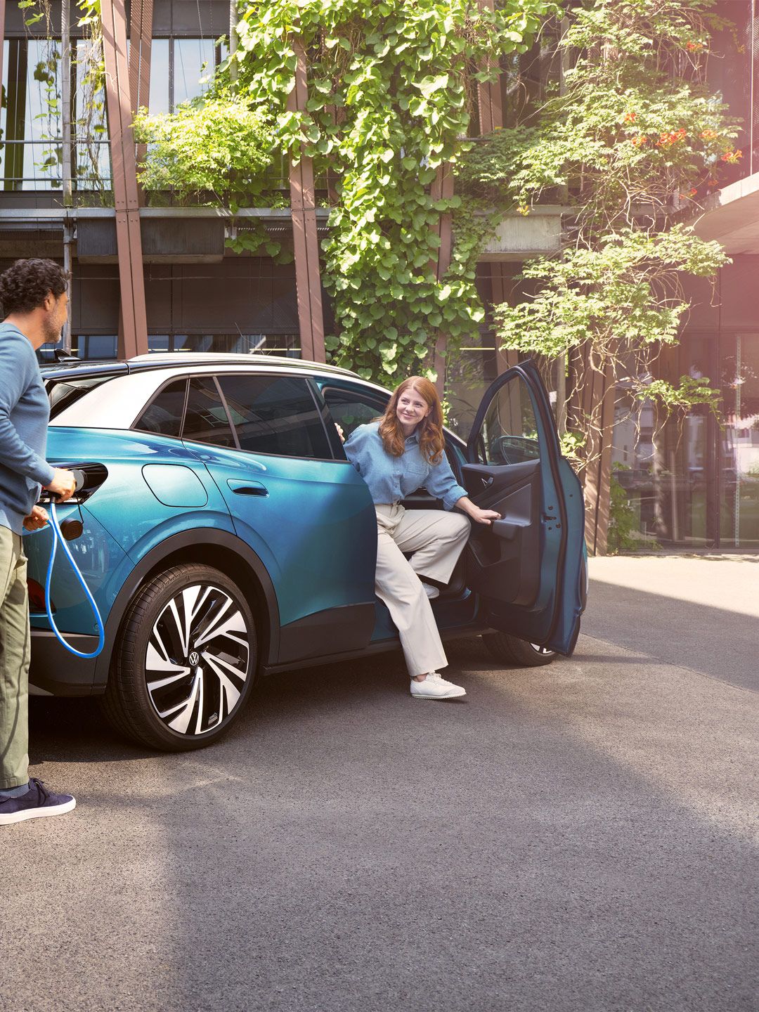 Ein Mann mit Ladekabel in der Hand geht zu einem blauen VW ID.4, der in einem begrünten Hof parkt.