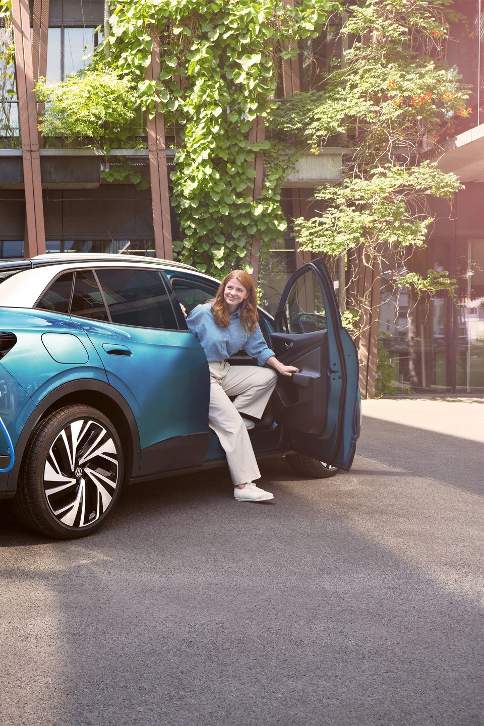 Ein Mann mit Ladekabel in der Hand geht zu einem blauen VW ID.4, der in einem begrünten Hof parkt.