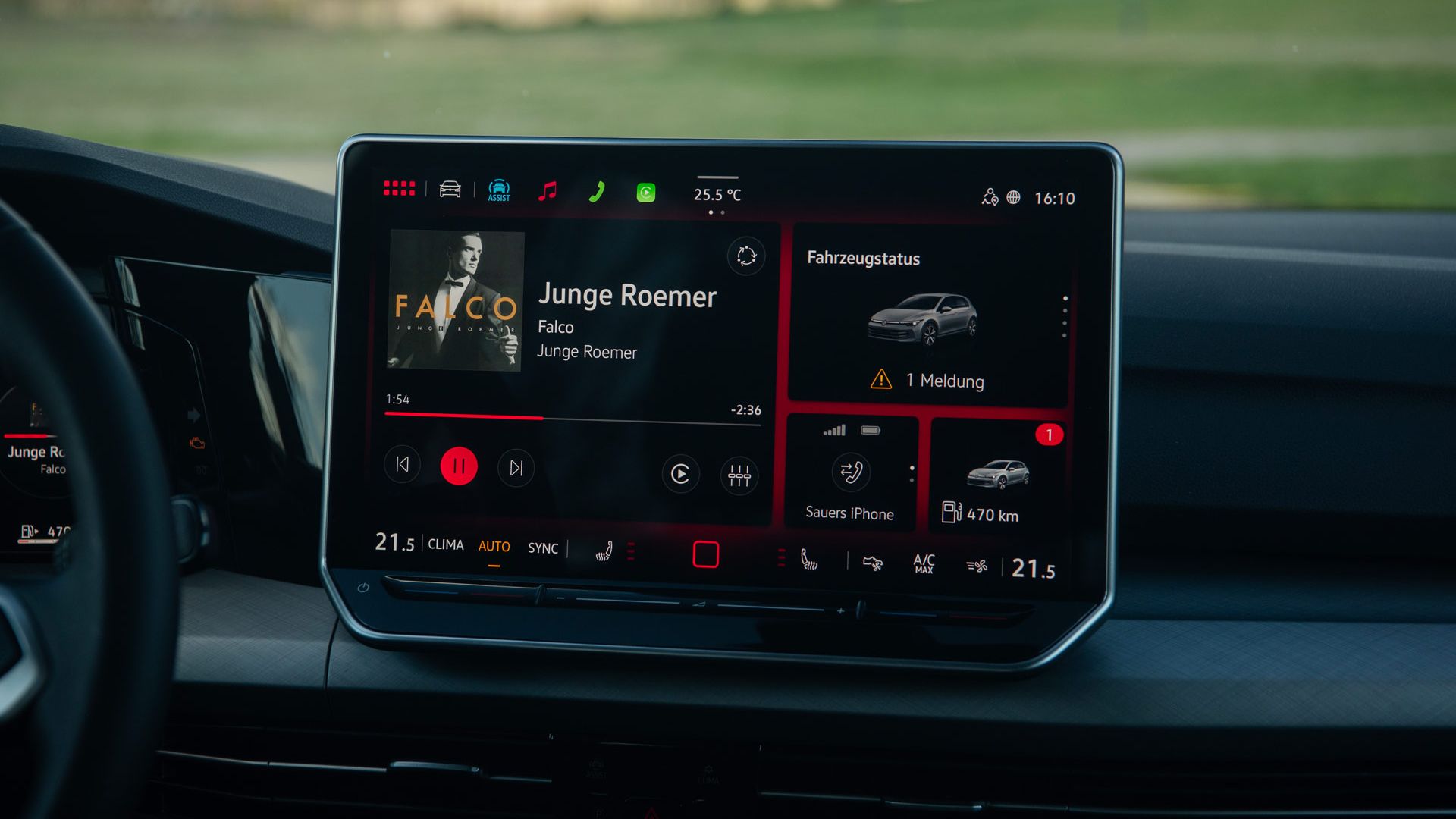 Zentral im Bild ist das Infotainment-Display eines Autos, auf dem Musik von Falco mit dem Titel „Junge Roemer“ abgespielt wird. Rechts auf dem Bildschirm werden der Fahrzeugstatus mit einer Meldung sowie Fahrdaten wie Reichweite und verbundenes iPhone angezeigt. Unten sind Klimaeinstellungen und weitere Touch-Bedienelemente sichtbar, der Rest des Armaturenbretts bleibt dezent im Hintergrund.