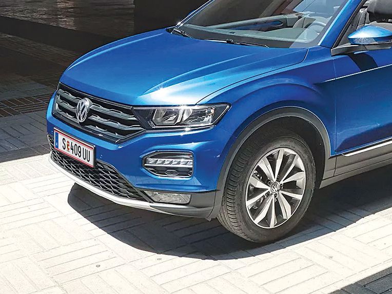 Ein blaues Volkswagen Cabriolet steht in einem sonnigen, überdachten Bereich. Es hat das Verdeck geöffnet. Die Umgebung ist modern und urban.
