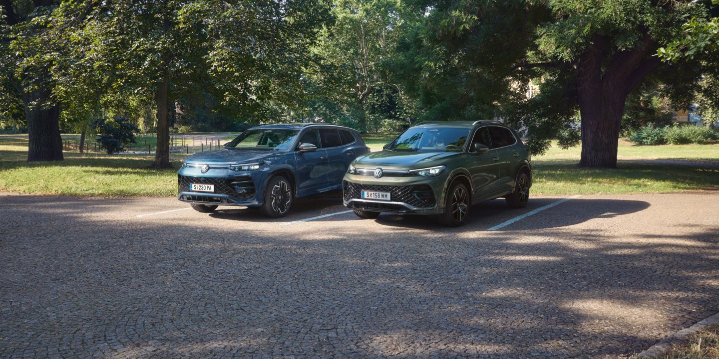 Blauer Tiguan R-Line Plug-In-Hybrid und grüner Tayron R-Line Plug-In- Hybrid parken nebeneinander im schattigen Parkbereich.