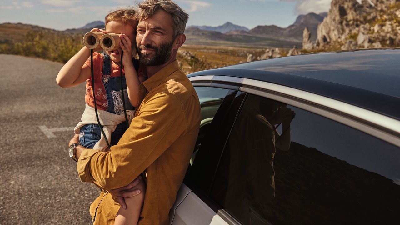 Ein Erwachsener lehnt an einem Volkswagen und hält ein Kind, das durch ein Fernglas schaut.