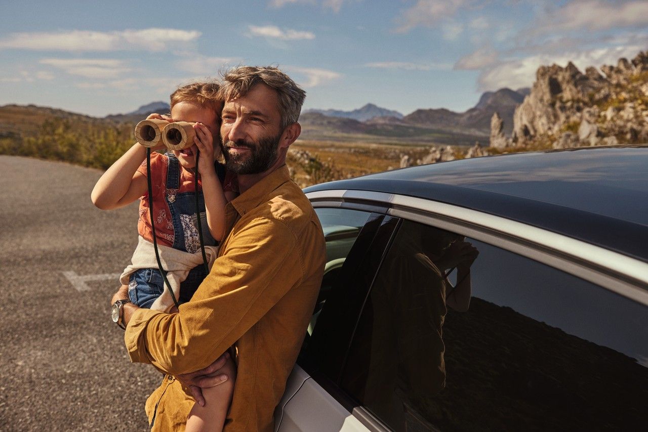 Ein Erwachsener lehnt an einem Volkswagen und hält ein Kind, das durch ein Fernglas schaut.
