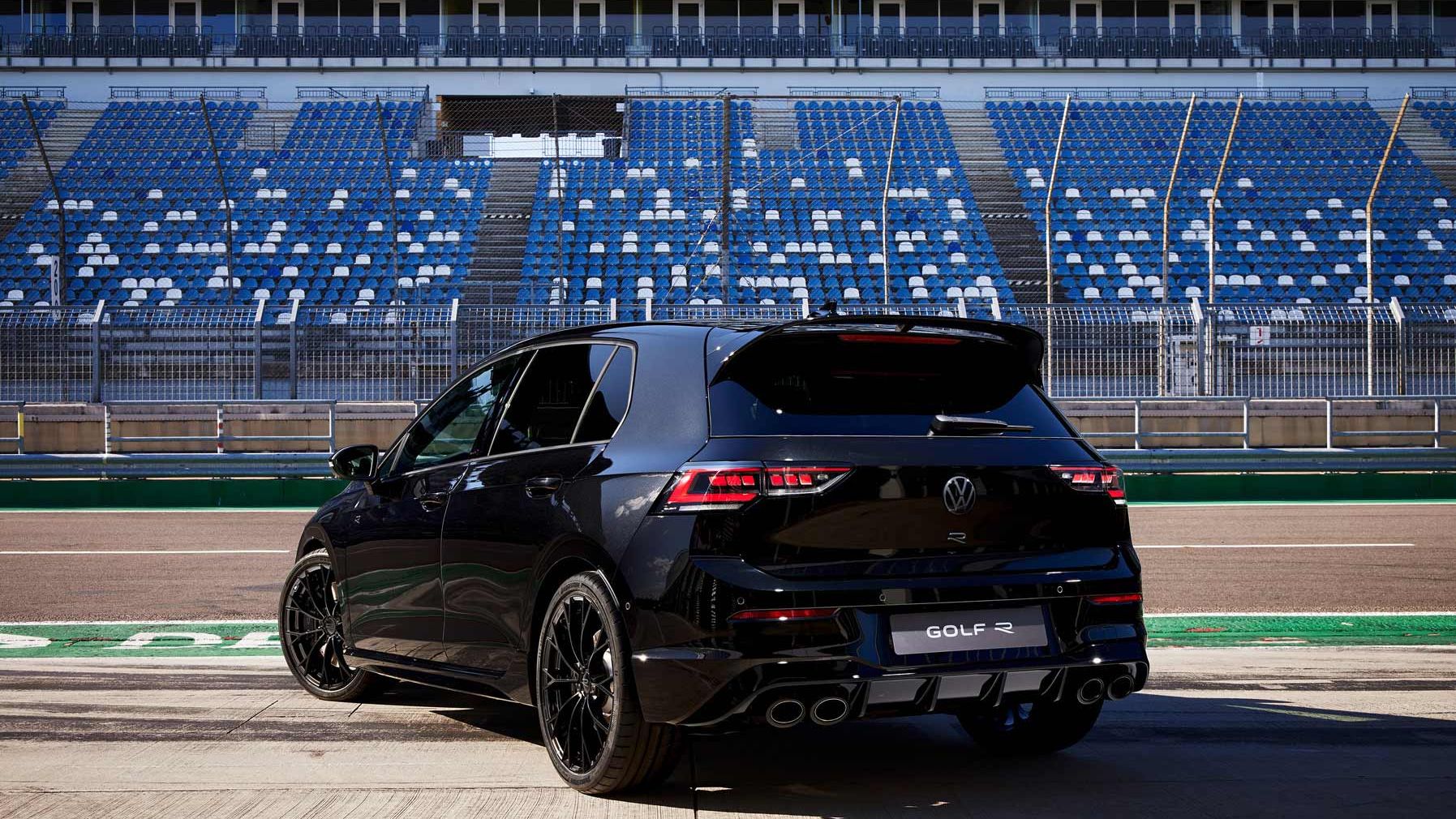 Ein schwarzer Volkswagen Golf R parkt auf einer Rennstrecke. Im Hintergrund sind leere Tribünen mit blauen und weißen Sitzen zu sehen.