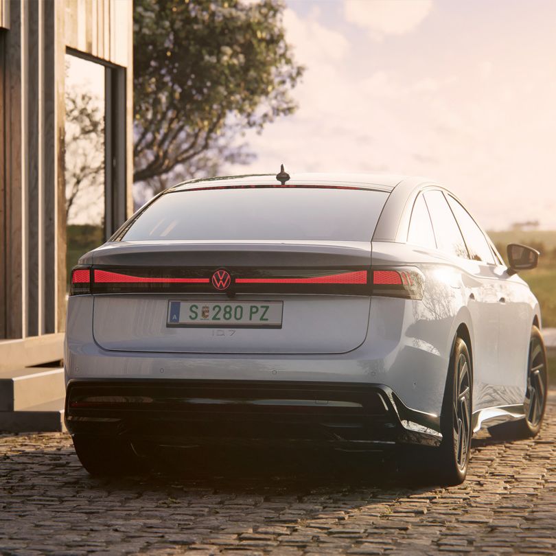 Heckansicht eines weißen VW ID.7, der neben einer Ladesäule steht. Die Heck-Lichtsignatur mit Volkswagen-Logo leuchtet rot.