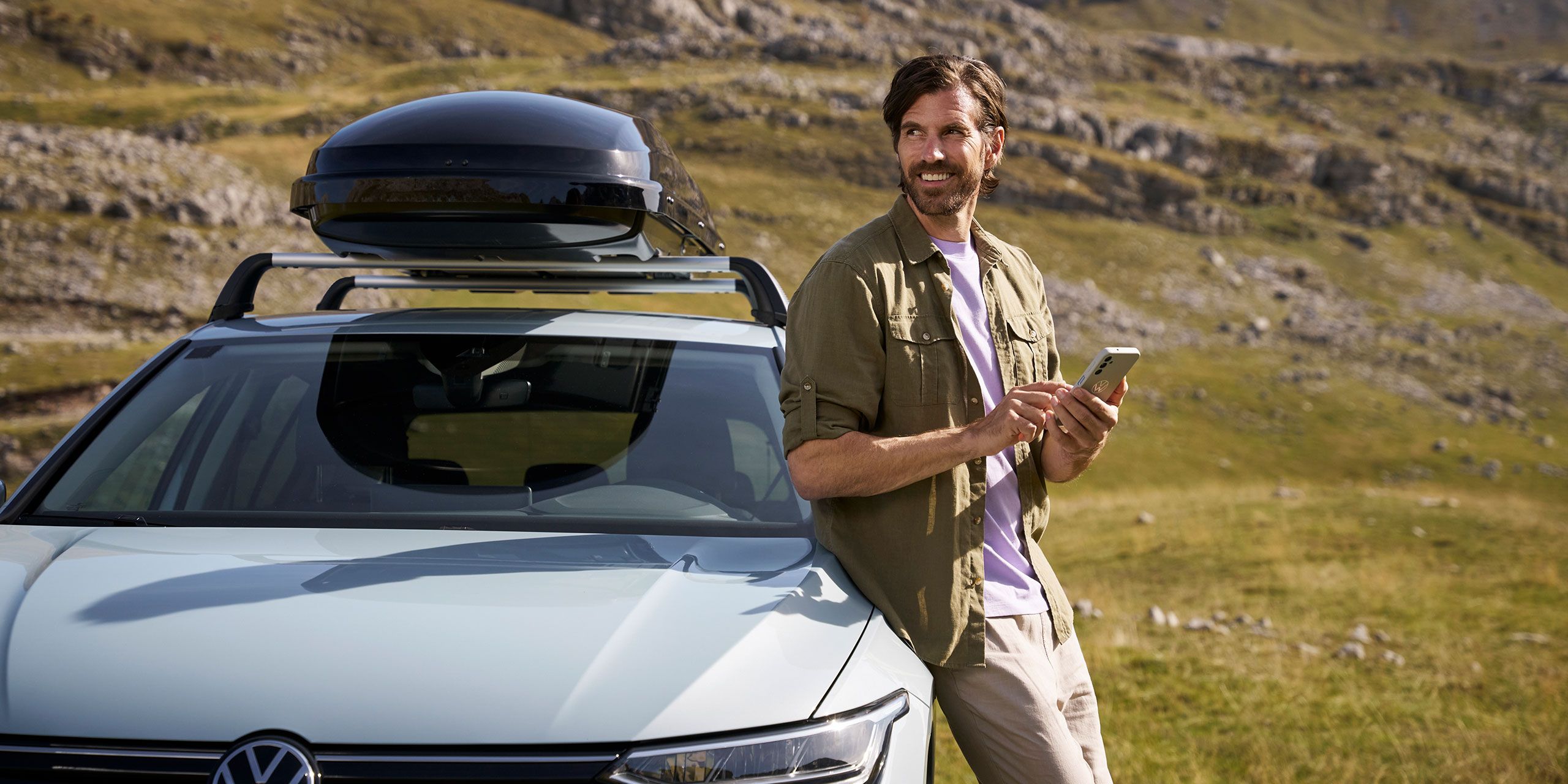 Ein Mann lehnt an einem silbernen VW Modell an und schaut in die Ferne, während er sein Handy in der Hand hält.