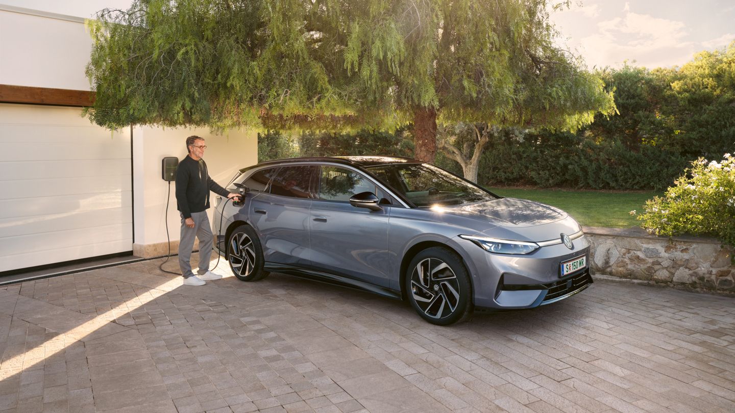 Ein Mann steht neben einem blauen VW ID.7 Tourer, der vor einer Garage an einer Wallbox lädt.