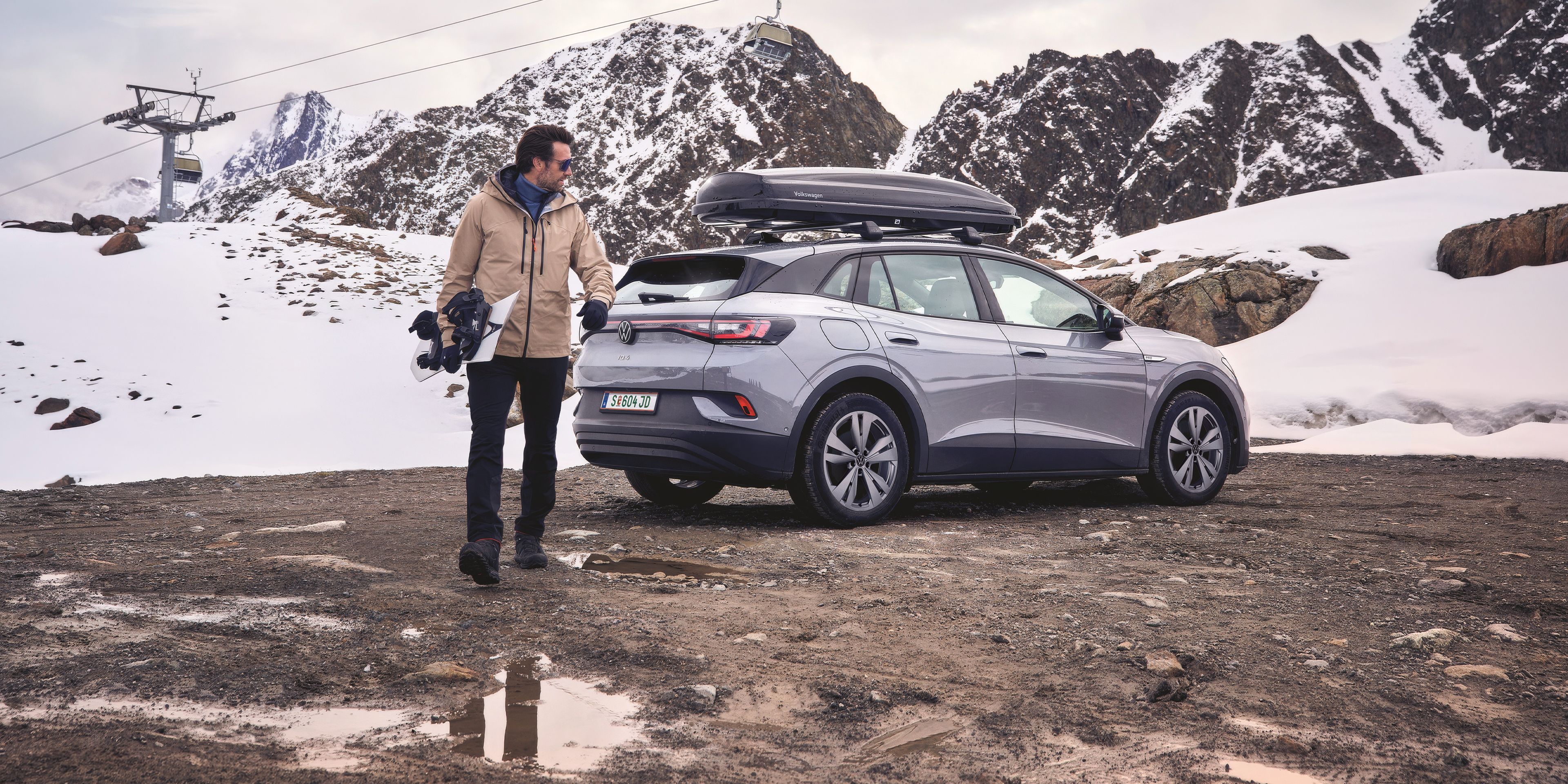 Ein Mann steht neben einem grauen Volkswagen SUV mit Dachbox. Er hält ein Snowboard in der Hand und trägt Winterkleidung, während der Boden matschig und teils von Pfützen bedeckt ist.