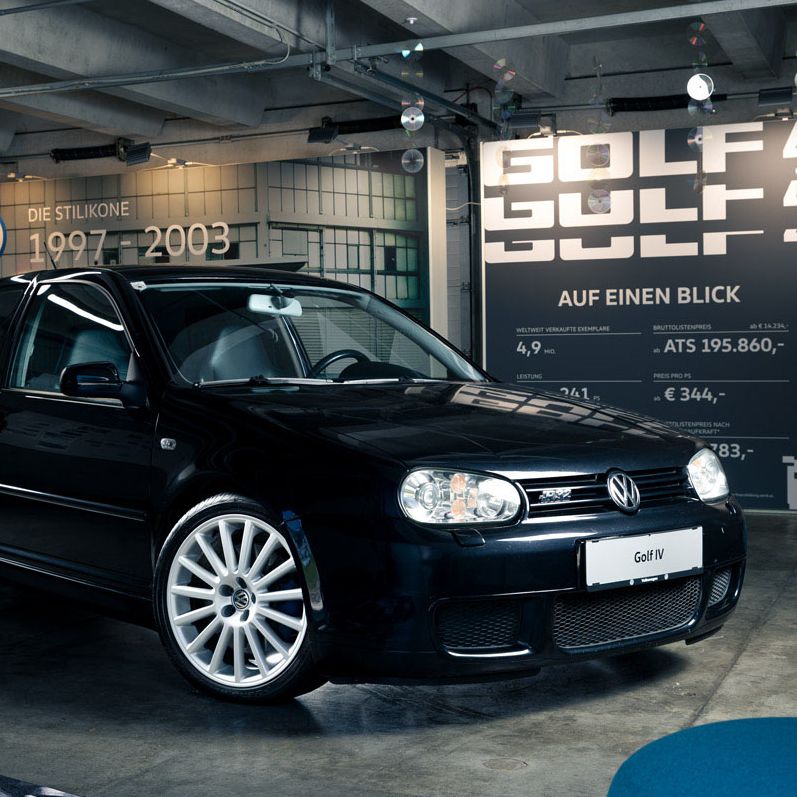 Ein schwarzer Volkswagen Golf IV steht in einer modern gestalteten Garage oder Ausstellung. Das Auto hat auffällige weiße Felgen und ist in einem glänzenden Zustand. Im Hintergrund sind Informationen über das Modell sowie Jahre der Produktion (1997-2003) an der Wand zu sehen.