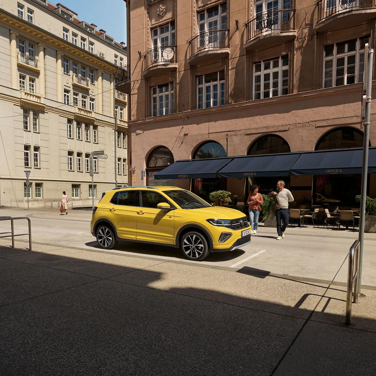 VW T-Cross in gelb parkt seitlich vor einem urbanen Straßencafé, ein Päarchen geht auf den Wagen zu.