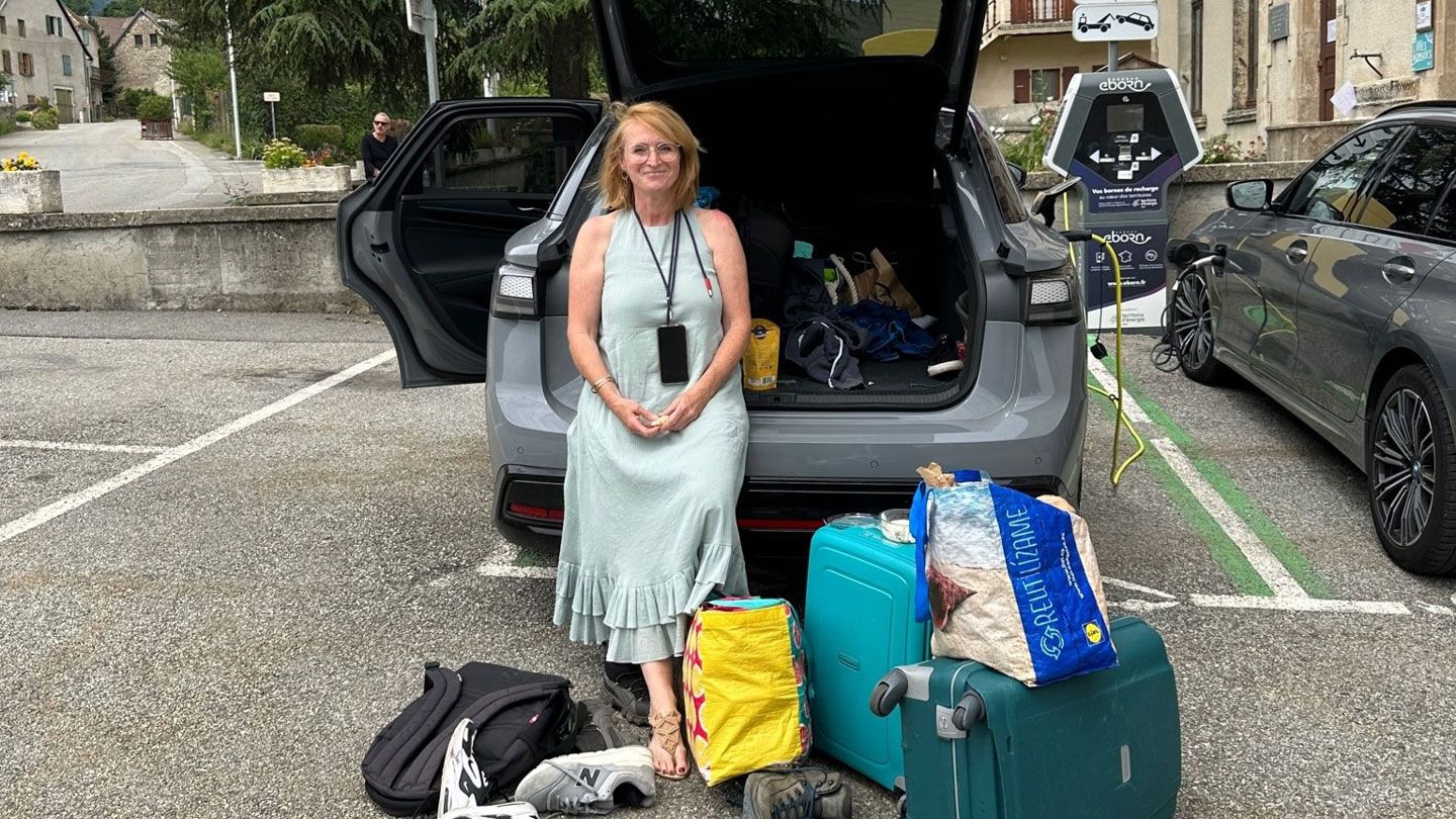 Eine Frau sitzt vor einem geparkten Auto, umgeben von Gepäckstücken wie Koffern und Taschen. Hinter ihr ist der geöffnete Kofferraum des Autos zu sehen. Im Hintergrund befinden sich Gebäude und eine Straße.