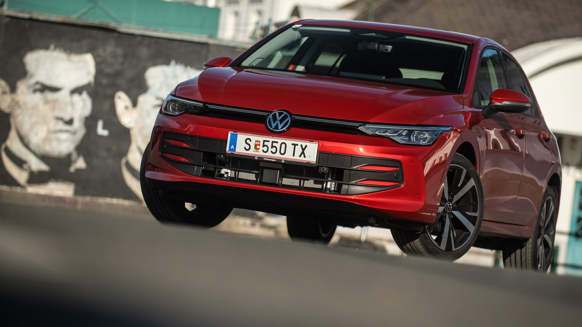 Roter Volkswagen Golf, schräg von vorne fotografiert, steht auf einer Straße vor einer Häuserkulisse.  
Das Auto wirkt dynamisch, als wäre es in Bewegung, und die Sonne erzeugt Reflexionen auf dem Lack.