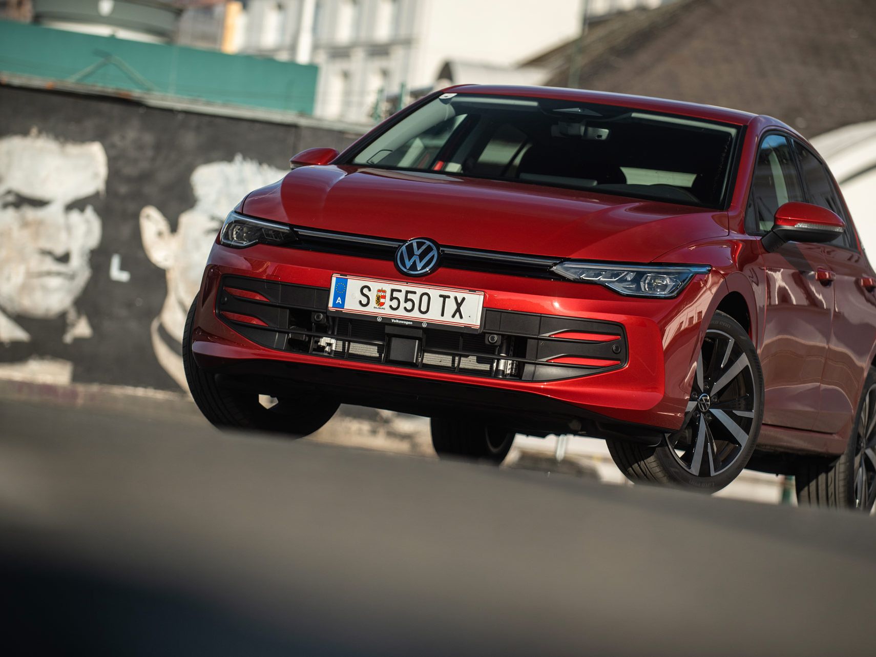 Roter Volkswagen Golf, schräg von vorne fotografiert, steht auf einer Straße vor einer Häuserkulisse.  
Das Auto wirkt dynamisch, als wäre es in Bewegung, und die Sonne erzeugt Reflexionen auf dem Lack.