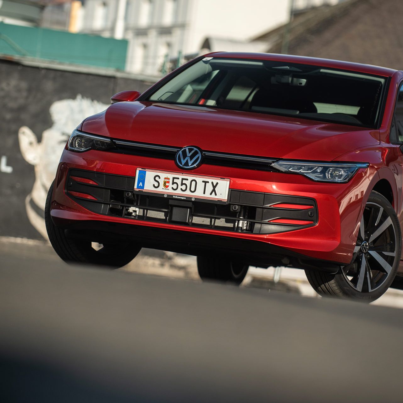 Roter Volkswagen Golf, schräg von vorne fotografiert, steht auf einer Straße vor einer Häuserkulisse.  
Das Auto wirkt dynamisch, als wäre es in Bewegung, und die Sonne erzeugt Reflexionen auf dem Lack.