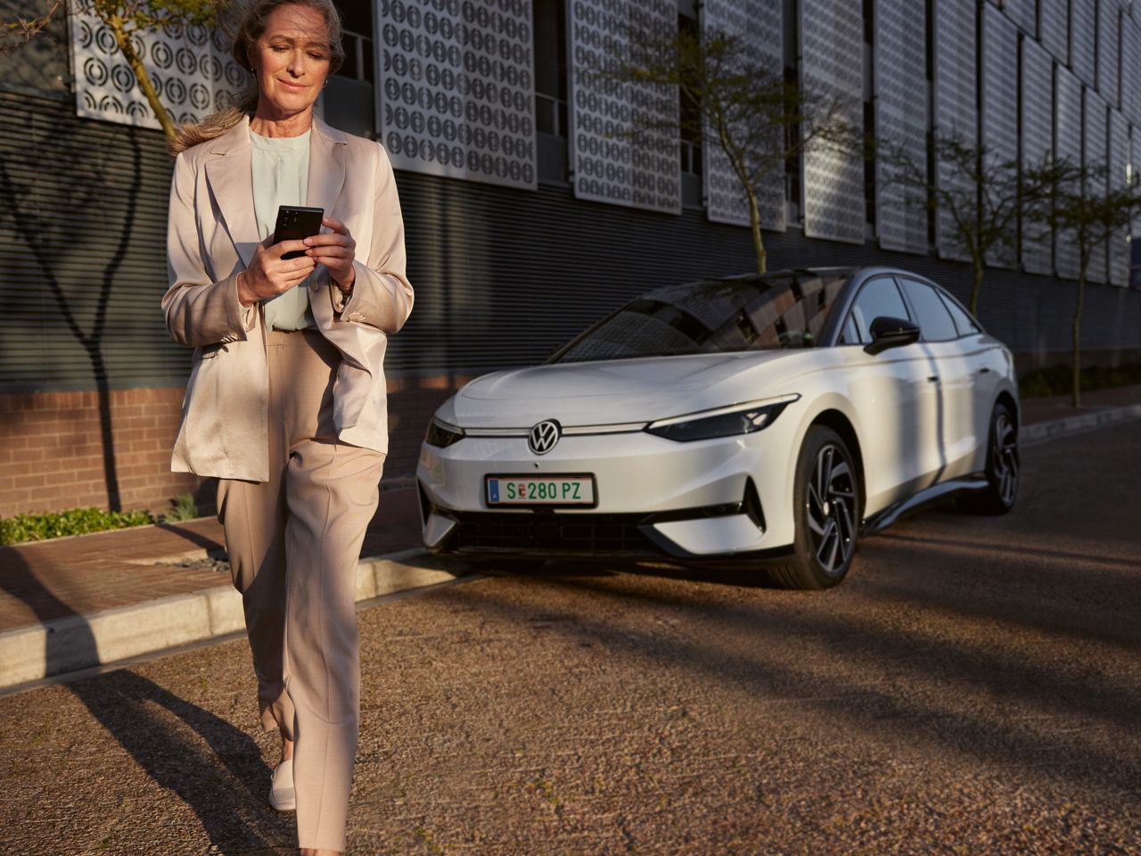 Eine Frau im Anzug schaut auf ihr Smartphone, im Hintergrund parkt ein weißer VW ID.7 am Straßenrand.
