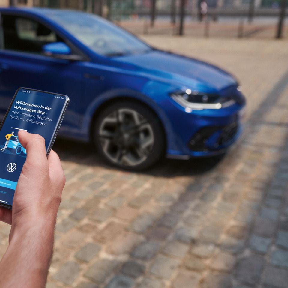 Eine Person hält ein Smartphone in der Hand mit der Volkswagen App, im Hintergrund steht ein blauer VW Polo R-Line.